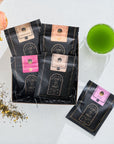 A display of five black packets of Green Magic Sampler Box from "Magic Hour" in various flavors, laid out on a white surface alongside a glass of vibrant green tea. Some loose leaf tea is scattered near the packets, and a bouquet of peach-colored flowers is partially visible.