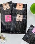 A display of five black packets of Green Magic Sampler Box from "Magic Hour" in various flavors, laid out on a white surface alongside a glass of vibrant green tea. Some loose leaf tea is scattered near the packets, and a bouquet of peach-colored flowers is partially visible.
