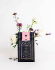 A black and pink package of "Gypsy Rose Black Tea" by Magic Hour stands on a white surface, adorned with the label "Time for Infinity". A few white and purple flowers, including lavender, are creatively arranged around and behind the package, giving it a fresh, elegant look.
