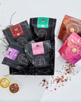 A box filled with assorted packages of loose leaf tea is displayed. The packages showcase various vibrant designs and colors, including black, pink, red, and green. A gold tea infuser with dried organic tea leaves is placed beside the **Magic Hour Herbal Magic Sampler Tea Box: Caffeine-Free Teas for Sleep, Digestion & Immunity** on a white surface.