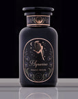 A stylish black bottle labeled "Magic Hour Astrology Bundle: Capricorn, Aquarius, Pisces" stands on a reflective surface. The label features an artistic depiction of a figure pouring water, surrounded by floral and star designs in gold. The cap of the bottle has a similar ornate design, hinting at the premium organic tea within.