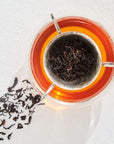 A glass cup filled with amber-colored Tea of The Rising Sun: Japanese Breakfast from Magic Hour sits on a white surface, with a metal tea infuser containing loose black tea leaves inside the cup. Loose leaf tea is scattered next to the cup, casting a shadow on the surface.