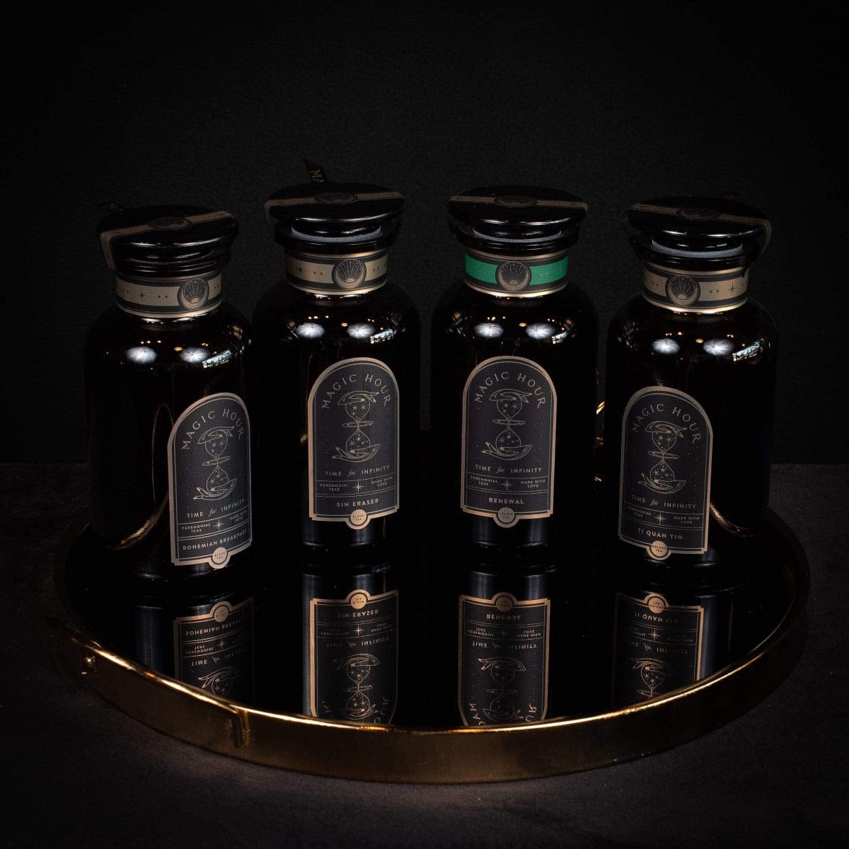 Four dark glass bottles with decorative labels stand on a golden tray against a dark background. The intricate designs read "Weight No More - Luxury Slimming Teas of the Far East," hinting at an elegant tea ceremony. One bottle, marked with a green label accent, suggests detoxifying qualities, while the others remain uniformly black.