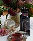 A colorful floral arrangement sits in a vase on a table next to a bottle labeled "Magic Hour" and a teacup filled with Soulmate: Chocolate-Raspberry-Rose Black Tea for Finding & Celebrating Love by Magic Hour. A glass orb and a single pink rose are also on the table, creating an elegant and inviting scene.