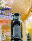 A visually appealing arrangement featuring a dark glass bottle of Magic Hour Renewal: Peach-Goji-Rose Oolong Tea placed on a reflective surface. It is flanked by two ornate glasses containing colorful beverages, with a bouquet of flowers and a decorative gold background enhancing the scene, highlighting the essence of loose leaf tea.