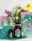 An arrangement of Queen of Kyoto Case Packs by Magic Hour, showcasing floral packaging inspired by cherry blossoms and Kyoto. The collection features jars, pouches, and boxes adorned with pink flowers. Highlighting the Japanese Cherry Blossom Festival, the display includes a bouquet of assorted flowers in a vase as the backdrop.