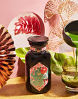 A vibrant setup featuring a jar of Queen of Kyoto Case Packs matcha powder by Magic Hour with an ornate label, surrounded by elegant decorations including a copper fan, tropical leaf, and abstract pink art. A hand holds a black teapot, pouring matcha tea into a clear glass on a gold surface, reminiscent of the Japanese Cherry Blossom Festival.