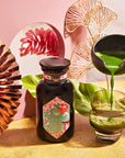 A vibrant setup featuring a jar of Queen of Kyoto Case Packs matcha powder by Magic Hour with an ornate label, surrounded by elegant decorations including a copper fan, tropical leaf, and abstract pink art. A hand holds a black teapot, pouring matcha tea into a clear glass on a gold surface, reminiscent of the Japanese Cherry Blossom Festival.