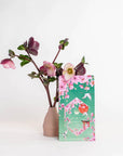 A Queen of Kyoto Case Pack from Magic Hour, a decorative light green bag adorned with pink flowers and birds, stands in front of a pink vase holding a branch with blooming pink flowers. The scene evokes the beauty of the Japanese Cherry Blossom Festival, and the plain white background highlights the vibrant and delicate details of these exquisite items.