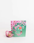 A green package adorned with pink floral designs and decorated with a transparent window revealing its contents. It is labeled with the "Magic Hour" brand and named "Queen of Kyoto Case Packs," featuring organic ingredients. A pink and white orchid flower sits beside the package against a plain white background.