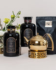 A display of tea products featuring a shiny gold teapot with a perforated stand, two black jars labeled "Big Magic: Violet Glass Apothecary Jar for Bulk Tea" by Magic Hour, and a floral arrangement with white flowers in the background. The setup is arranged on a light-colored surface, complemented by an enticingly black package of loose leaf tea.