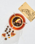A clear glass cup filled with organic tea, a metal infuser with dried tea balls, and a slice of dried orange rest on a white surface. Beside the cup, there is a "Strength" tarot card depicting a woman holding a lion. Dried oranges and loose leaf tea from The Astrology Collection - Luxe Sampler Sips by Magic Hour are scattered around.