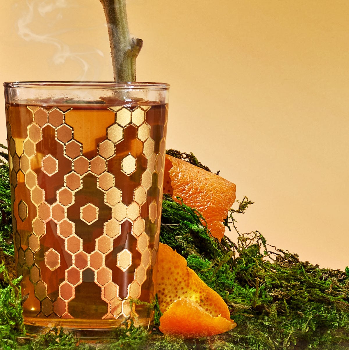 A clear glass filled with steaming hot detox tea sits atop a bed of green moss, showcasing an elegant honeycomb pattern in gold. Orange peel pieces are scattered around the moss, lending a vibrant touch to the earthy scene, reminiscent of the exotic offerings in the Spice Lovers Starter Set.