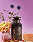 A pink and purple-toned still life image featuring a black bottle labeled "Magic Hour," adorned with intricate gold designs. Surrounding it are vibrant flowers, a glass teacup with steaming Libra- Pistachio-Rose Persian Love Cake Tea with White Pearls & Shatavari, dried herbs, and purple thistle-like flowers.
