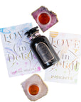 A bottle of Magic Hour tea is surrounded by two copies of "Love in Detail By Zhena Muzyka," which offer spiritual guidance from a women’s empowerment coach, and three teacups with a red beverage. The books feature decorative covers, and the teacups rest on jagged-edged coasters.