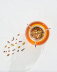 A cup of organic tea with an infuser containing loose leaf tea is viewed from above. The tea is a vibrant orange color, and the infuser reveals an assortment of leaves and spices. Some loose tea leaves are scattered on the white surface next to the cup, capturing a perfect Magic Hour Classics - Luxe Sampler Sips moment.