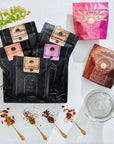 A display of various packaged tea blends, including Magic Hour's The Best of Magic Hour Sampler Set with Tea in Hand Teapot, features five black and two colored packets. The black packets have copper labels. Beside the packages are three gold spoons with loose leaf tea, a clear glass teapot, and green foliage providing a decorative touch.