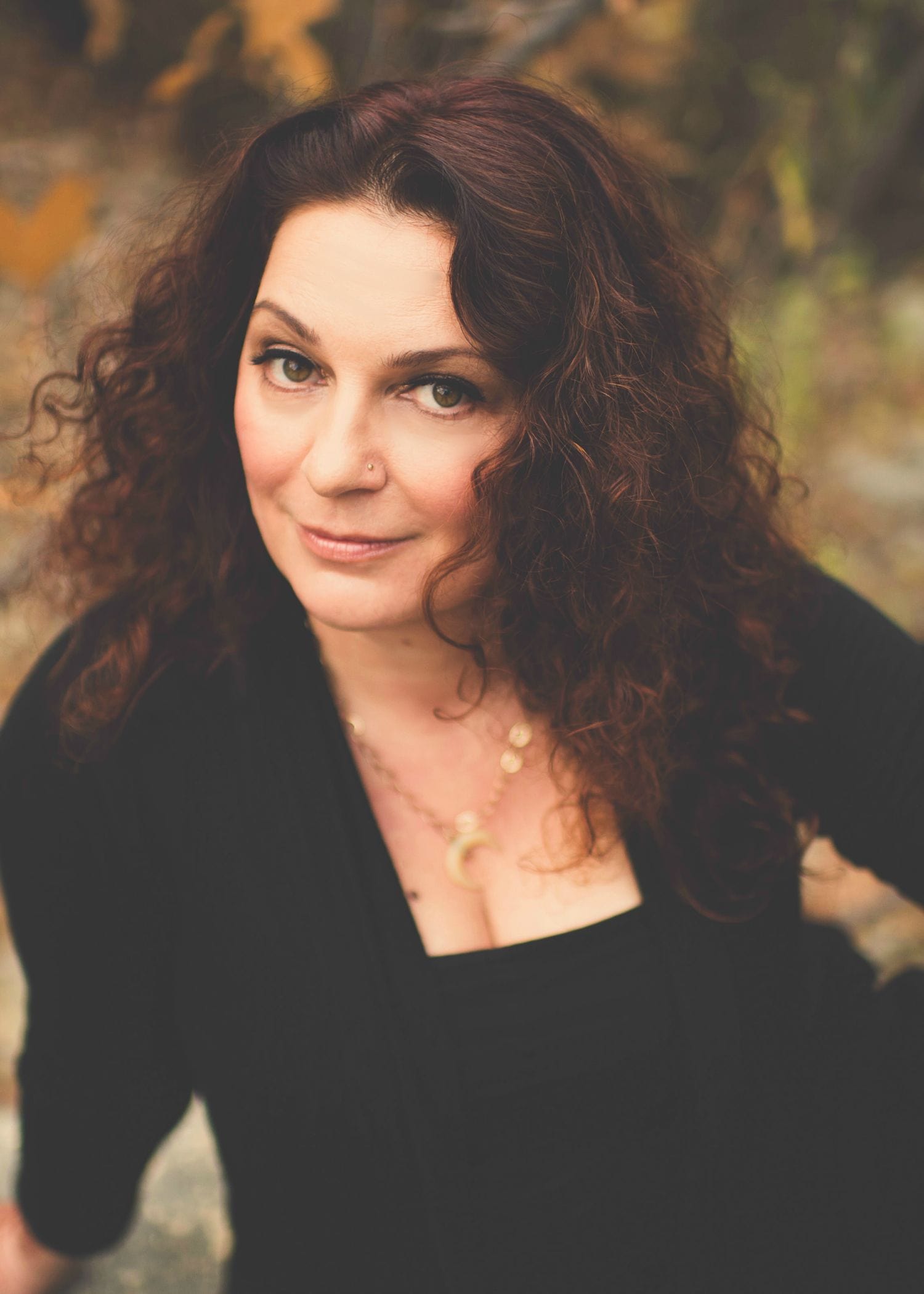 A person with long, curly brown hair and a nose piercing is looking up at the camera with a slight smile. They are wearing a black top and necklace and are outdoors, surrounded by greenery. Inspired by the Love & Intimacy Box with Raspberry Earl White, their peaceful aura radiates serenity.