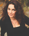 A person with long, curly brown hair and a nose piercing is looking up at the camera with a slight smile. They are wearing a black top and necklace and are outdoors, surrounded by greenery. Inspired by the Love & Intimacy Box with Raspberry Earl White, their peaceful aura radiates serenity.
