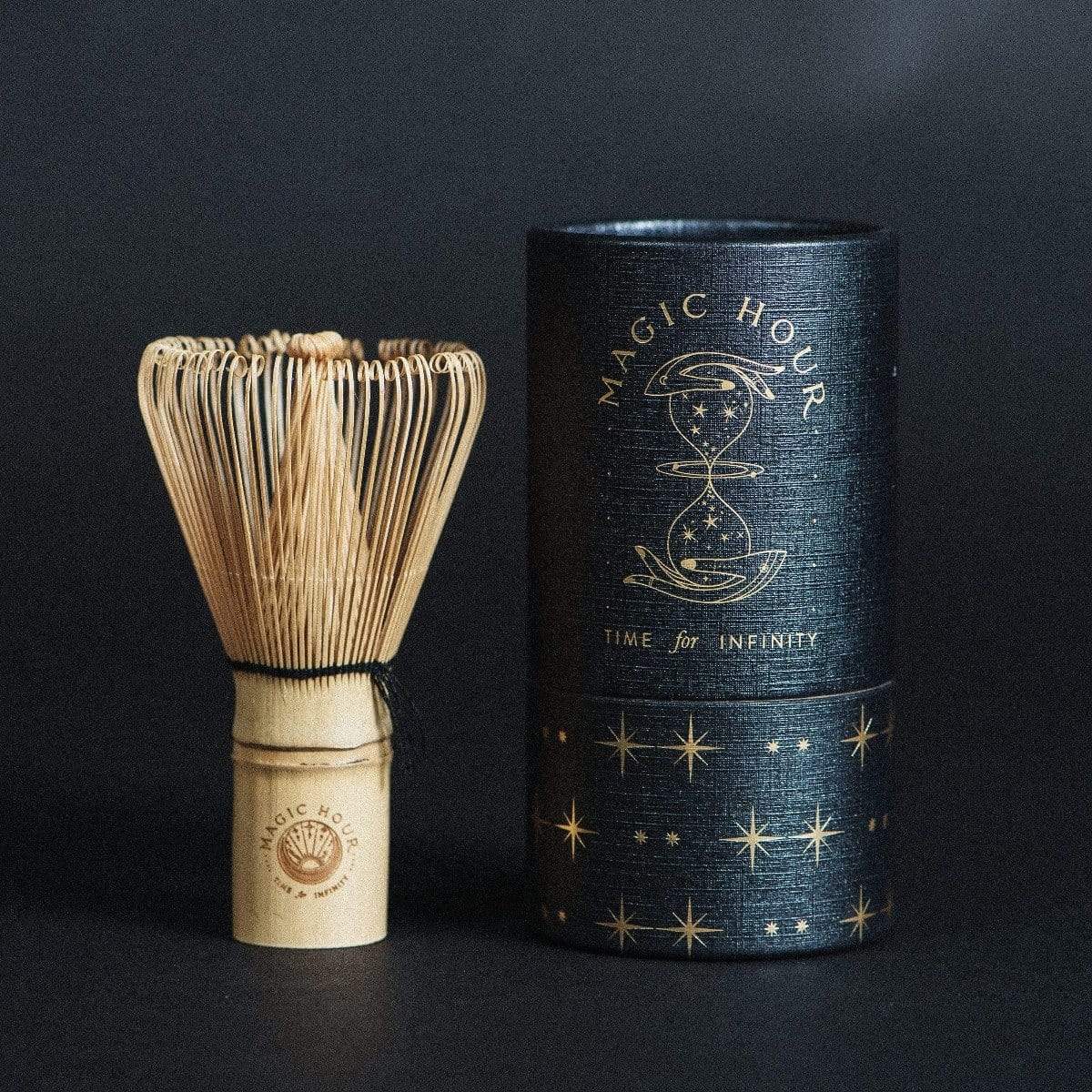 A Ceremonial Matcha Traveler Gift Box with fine, thin prongs stands next to a cylindrical black container. The container features gold text reading "MAGIC HOUR" with an hourglass illustration and celestial patterns. The words "TIME for INFINITY" are written below the hourglass, making it perfect for savoring your Magic Hour Tea moments.