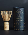 A traditional bamboo tea whisk is placed beside a decorative black cylindrical container with the words "Magic Hour - Matcha 1111 : Gift Set" and starry illustrations. Perfect for brewing loose leaf or organic tea, both items feature a matching logo. The background is dark.