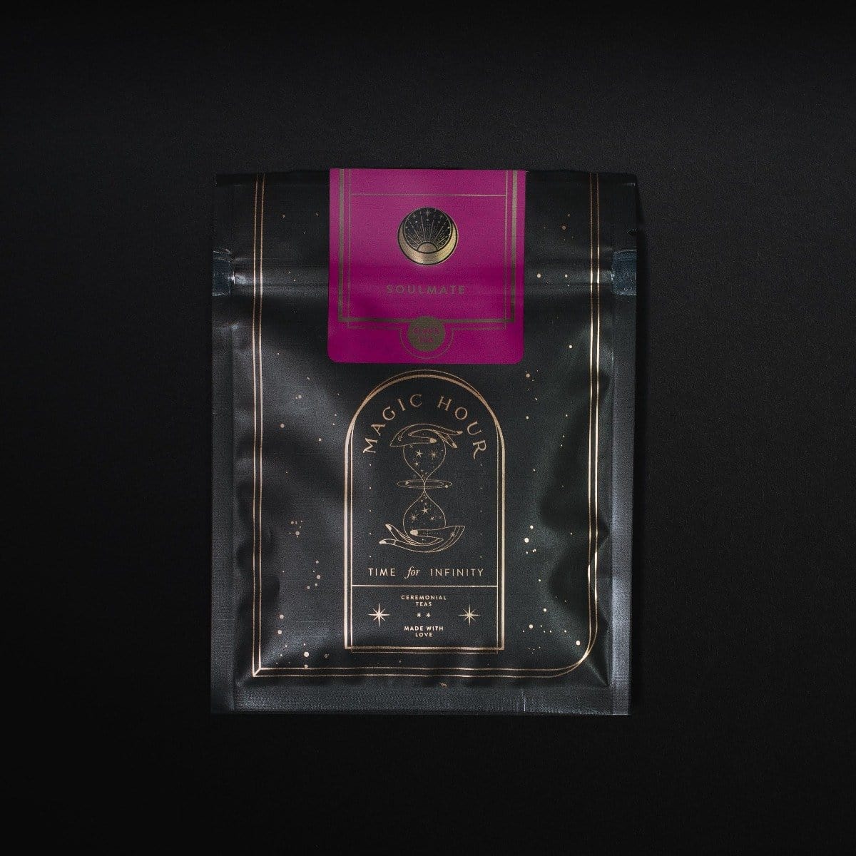 A black-colored Magic Hour Tea package is shown against a dark background. The bag has a decorative gold design with an hourglass illustration, and it reads "Time for Infinity." A pink label with a circular emblem that says "Soulmate: Chocolate-Raspberry-Rose Black Tea for Finding & Celebrating Love" is attached at the top, highlighting its organic loose leaf tea inside.