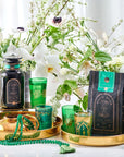 A luxurious tea setup features a black canister of Mantra Mint™ Herbal Tea from Club Magic Hour and a matching bag displayed on a golden tray. Green glasses adorned with golden patterns, a teapot, and a green beaded necklace complement the arrangement, surrounded by white flowers.