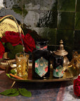 A decorative scene featuring a dark bottle with intricate golden and colorful designs, accompanied by a matching smaller container on an ornate golden tray showcases the opulence of Moonlight in Marrakesh: Spiced Mint Green Tea Case Packs by Magic Hour. Two lavishly adorned glasses are being filled with this exquisite Moroccan mint tea, while red roses and a teapot accentuate the setting.