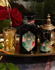 A decorative scene featuring a dark bottle with intricate golden and colorful designs, accompanied by a matching smaller container on an ornate golden tray showcases the opulence of Moonlight in Marrakesh: Spiced Mint Green Tea Case Packs by Magic Hour. Two lavishly adorned glasses are being filled with this exquisite Moroccan mint tea, while red roses and a teapot accentuate the setting.