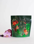 A stand-up pouch in a vibrant shade of green, adorned with intricate floral and animal designs, including flowers and a geometric pattern, is set against a clean white backdrop. Beside the pouch lies a delicate arrangement of pink and purple flowers, capturing the essence of Moroccan mint tea with subtle hints of organic ingredients. The pouch features "Moonlight in Marrakesh: Spiced Mint Green Tea Case Packs" from the brand Magic Hour.
