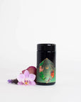 A Moonlight in Marrakesh: Spiced Mint Green Tea case pack by Magic Hour, with its black cylindrical container and decorative green and gold design, stands on a white surface. Surrounding it are pink and purple orchids, lavender flowers, and hints of Moroccan mint tea, adding a touch of nature and color to the scene.