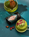 A top-down view of a matcha tea setup on a teal surface. It features a jar of Organic Ceremonial Matcha 700, a matcha whisk in a bowl of green tea, and a cup of Club Magic Hour Tea topped with rose petals and matcha powder. Loose rose petals are scattered around, adding an enchanting touch.