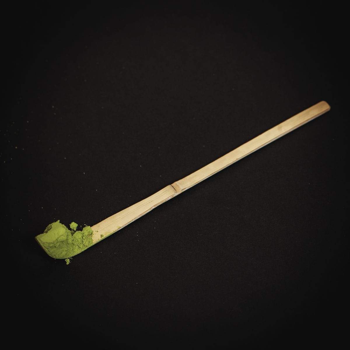 A bamboo tea scoop holds a small amount of Ceremonial Matcha Traveler Gift Box by Magic Hour against a dark, solid background. The scoop is simple and elongated, and the vivid green of the matcha contrasts sharply with the dark background.