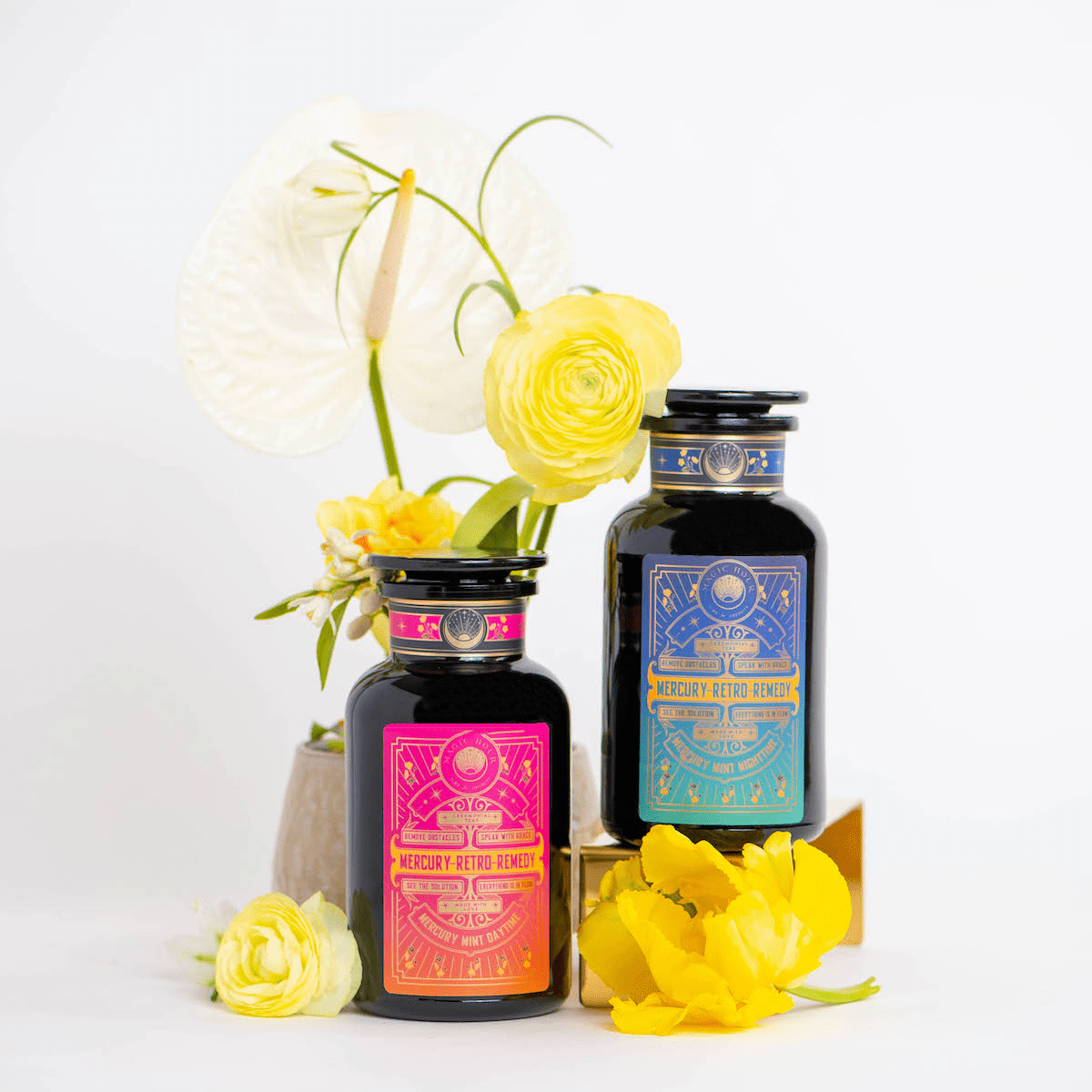 Two dark glass bottles with vibrant, ornate labels reading "Mercury Retro Remedy Bundle: Save 10% when you Buy the Bundle!" are displayed against a white background. The left bottle has a pink label, and the right bottle has a blue label. Bright yellow flowers surround the bottles, adding a decorative touch to these Magic Hour blends.