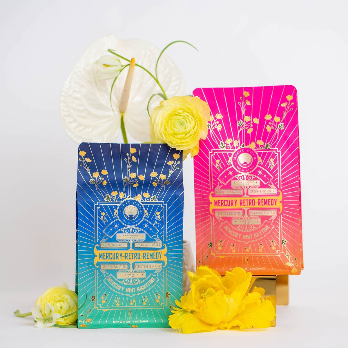 Two vibrantly colored Mercury Retro Remedy Bundle packages stand against a white background. The package on the left is blue and green, while the one on the right is pink and orange. Yellow flowers and white decorative leaves are arranged around the products, highlighting these enchanting Magic Hour blends. Save 10% when you buy the bundle!