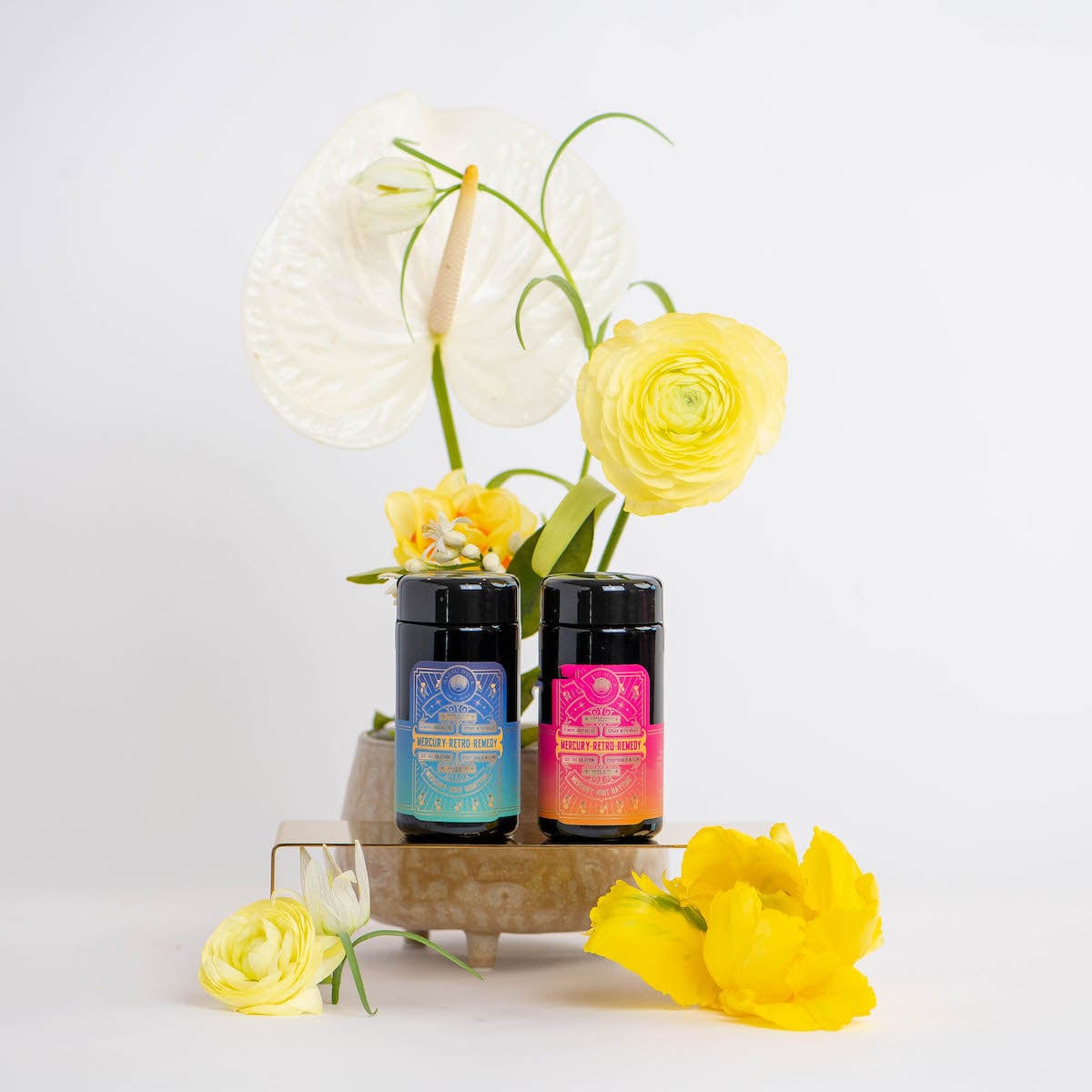 Two jars of colorful seasoning are displayed on a wooden stand, surrounded by vibrant yellow flowers and a white anthurium. The jars have intricate labels, adding a decorative touch to the bright and inviting arrangement—much like the Magic Hour's Mercury Retro Remedy Bundle: Save 10% when you Buy the Bundle!