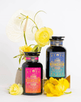 Two dark glass bottles with vibrant, ornate labels reading "Mercury Retro Remedy Bundle: Save 10% when you Buy the Bundle!" are displayed against a white background. The left bottle has a pink label, and the right bottle has a blue label. Bright yellow flowers surround the bottles, adding a decorative touch to these Magic Hour blends.