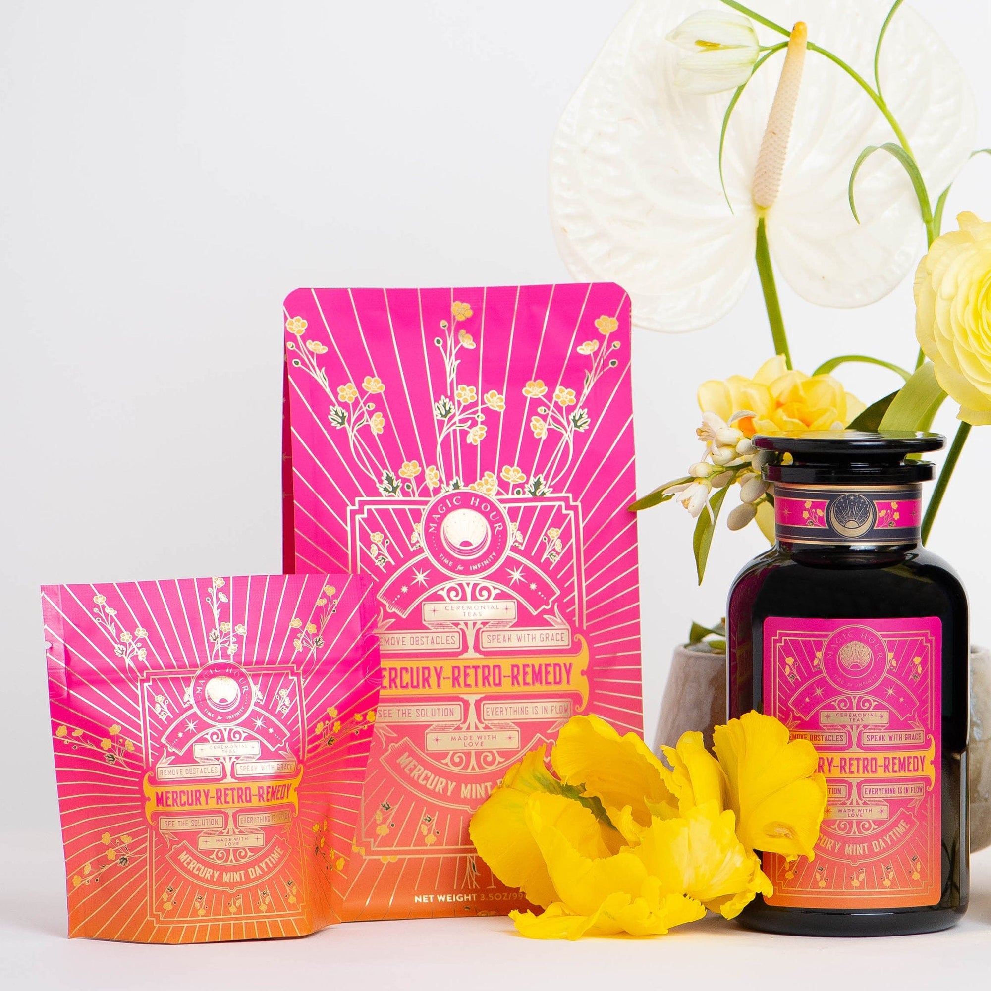 A vibrant display showcasing three Mercury Mint Daytime products in colorful pink and orange packaging adorned with astronomical designs. The set includes two pouches and a bottle of Vanilla Mint Lavender Jasmine Black Tea. They are surrounded by white and yellow flowers against a light background.