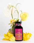 A black bottle with a vibrant pink and orange label reading "Mercury Mint Daytime: Vanilla Mint Lavender Jasmine Black Tea" is showcased in front of a white background. Surrounding the bottle are various flowers, including white, yellow, and green blooms, along with sprigs of lavender flowers, adding a botanical and fresh touch to the display.