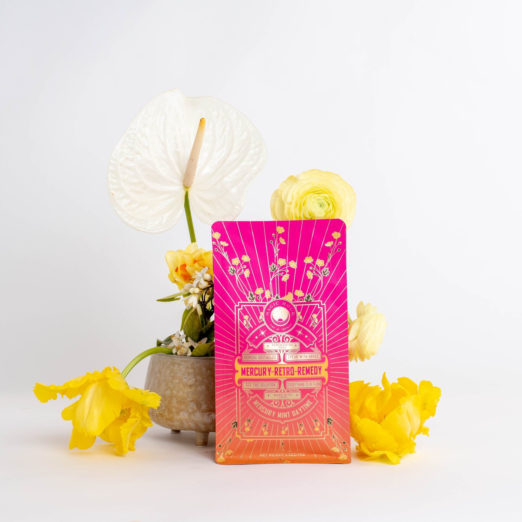 A vibrant pink and orange package labeled "Mercury Mint Daytime : Vanilla Mint Lavender Jasmine Black Tea" stands in front of a small vase containing white and yellow flowers. The backdrop is plain white, making the colors on the package and flowers pop, while hinting at the soothing presence of Organic Biodynamic Black Tea.