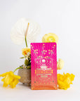 A vibrant pink and orange package labeled "Mercury Mint Daytime : Vanilla Mint Lavender Jasmine Black Tea" stands in front of a small vase containing white and yellow flowers. The backdrop is plain white, making the colors on the package and flowers pop, while hinting at the soothing presence of Organic Biodynamic Black Tea.