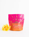 A vibrantly colored pouch labeled "Mercury Mint Daytime : Vanilla Mint Lavender Jasmine Black Tea" with decorative golden designs stands next to a yellow flower against a plain white background. The pouch, containing Organic Biodynamic Black Tea, transitions from pink at the top to orange at the bottom.