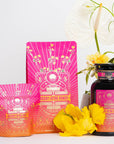 A vibrant display showcasing three Mercury Mint Daytime products in colorful pink and orange packaging adorned with astronomical designs. The set includes two pouches and a bottle of Vanilla Mint Lavender Jasmine Black Tea. They are surrounded by white and yellow flowers against a light background.