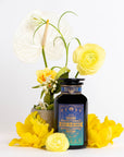 A black bottle with a blue and gold label reads "Mercury Mint Nighttime: Vanilla Mint Lavender Jasmine Tea- Caffeine Free!" The caffeine-free herbal blend is surrounded by an arrangement of yellow flowers and a white anthurium in a small pot. The backdrop is white, highlighting the colors of the flowers and the decorative bottle.