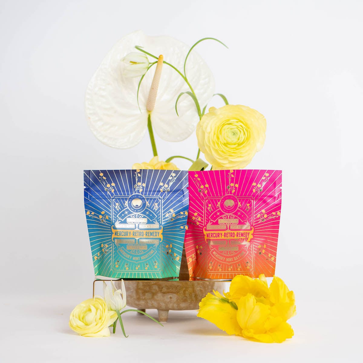 Two vibrantly colored packets—one blue, one pink—of "Mercury Retro Remedy Bundle: Save 10% when you Buy the Bundle!" herbal tea are displayed amidst bright yellow flowers and large white leaves, all set against a clean, white background. The packets feature celestial design elements from the Magic Hour blends collection.