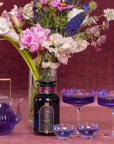 A vibrant setup featuring a dark bottle labeled "Symbeeosis: Beautifying Immunitea for the Queen Bee" from Magic Hour, a purple teapot of loose leaf tea, two purple-tinted glasses with a floral garnish, and a vase of diverse flowers including white lilies and pink peonies, arranged against a rich, burgundy background.