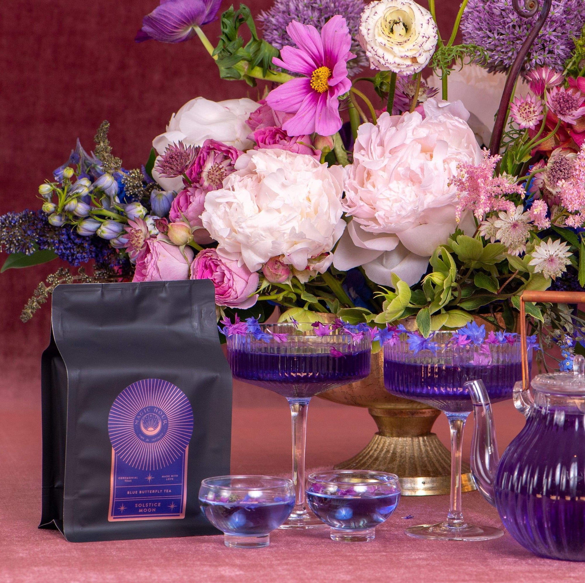 A dark bag of Solstice Moon™ coffee with a colorful label sits on a table surrounded by vibrant flowers in various shades of pink and purple. Nearby are elegant purple cocktails in glassware, a clear teapot filled with an organic tea blend, and whispers of Winter Solstice magic.