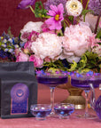 A dark bag of Solstice Moon™ coffee with a colorful label sits on a table surrounded by vibrant flowers in various shades of pink and purple. Nearby are elegant purple cocktails in glassware, a clear teapot filled with an organic tea blend, and whispers of Winter Solstice magic.