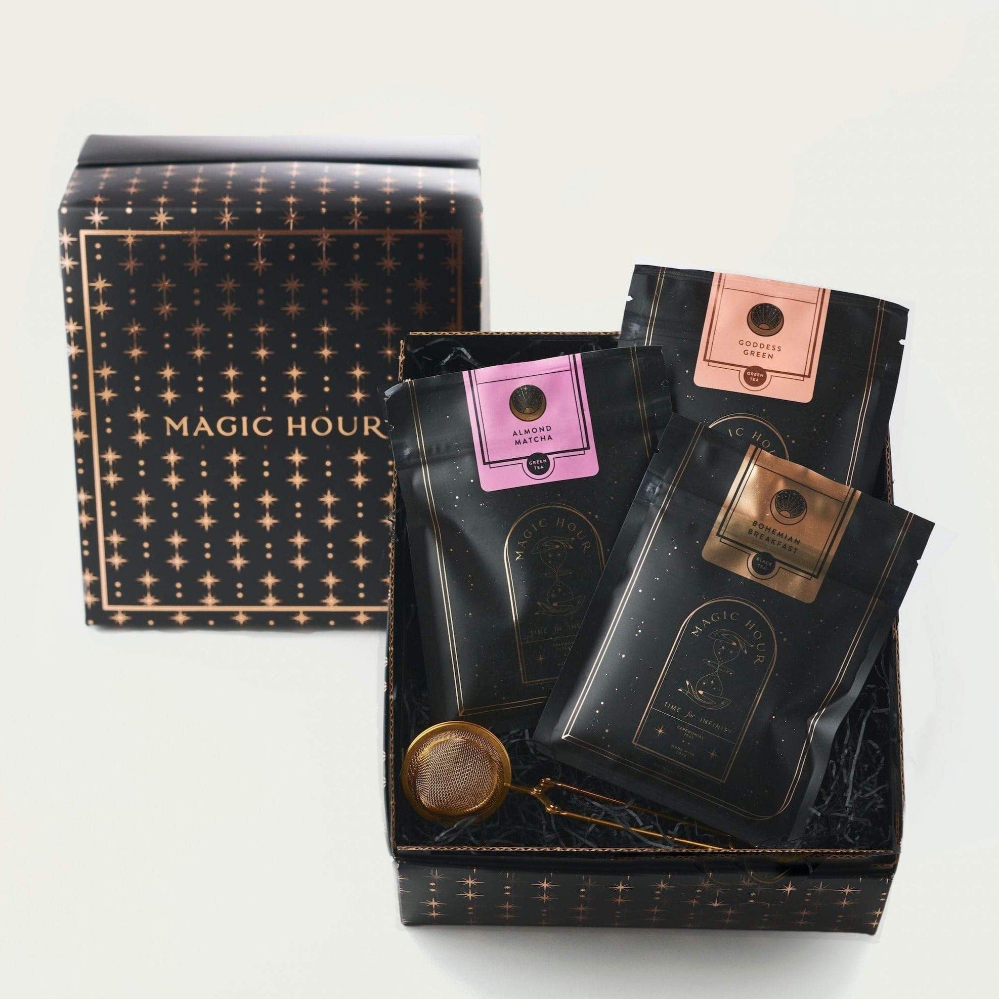 A gift box labeled "Sip Magic: Signature Blends of Magic Hour Ritual Gift" by Magic Hour contains three packages of black loose leaf tea with colorful labels: pink, brown, and copper. A tea infuser is also included. The packaging, adorned with a gold star pattern, adds an enchanting touch against the plain white background.