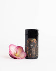 A sleek black cylindrical tea container from Magic Hour's Black Onyx - Chaga Maca Toffee Elixir Case Packs, featuring an intricate gold design, stands on a white background. Beside it rests a single pink and white flower, adding a touch of softness and color to the minimalist scene. This elegant presentation subtly suggests the benefits of caffeine-free, immunity-boosting teas like Chaga.